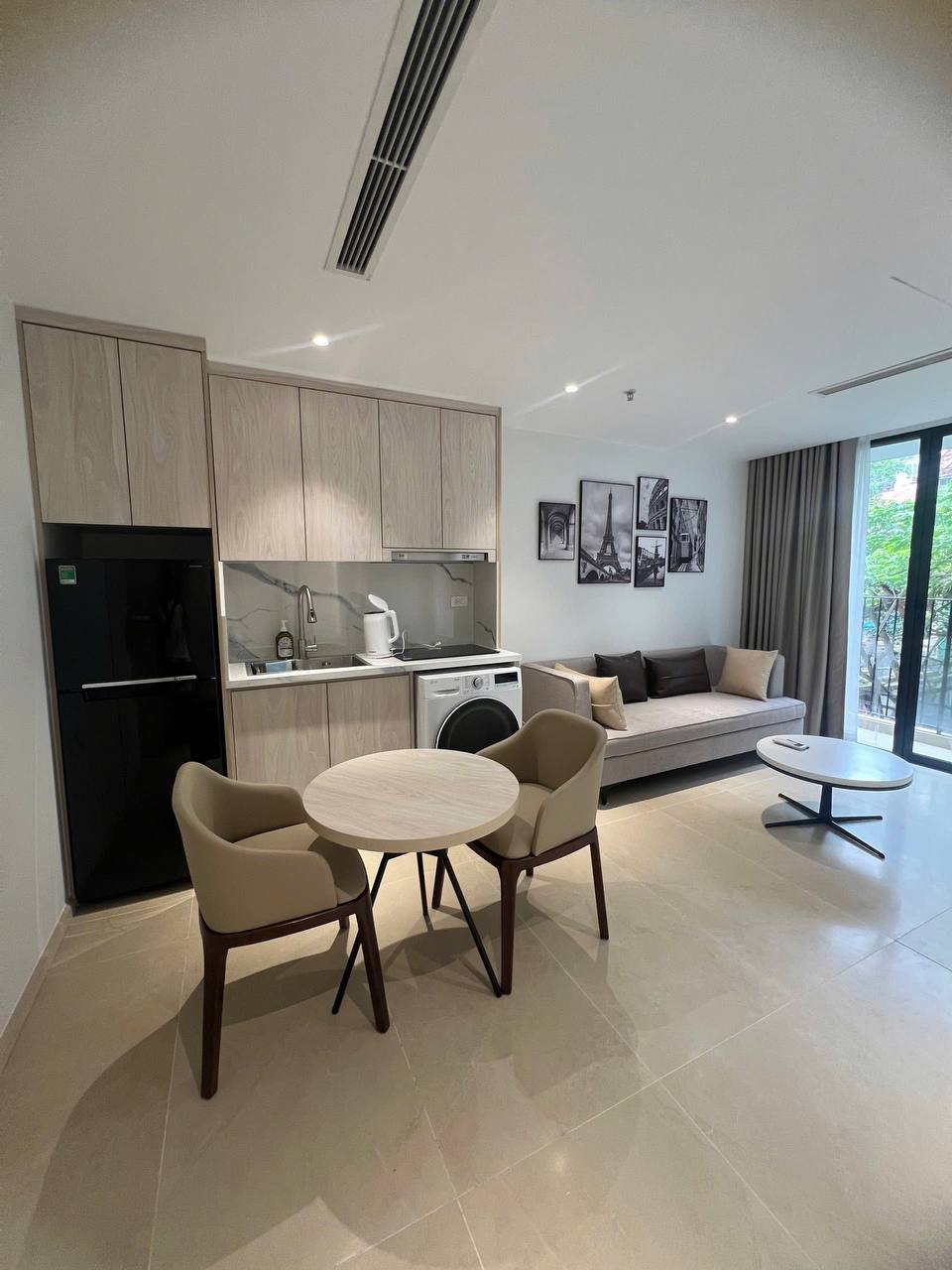 Phòng khách căn 1BR toàn cảnh - sofa xám, bàn trắng, tranh xe vàng Paris, cửa kính